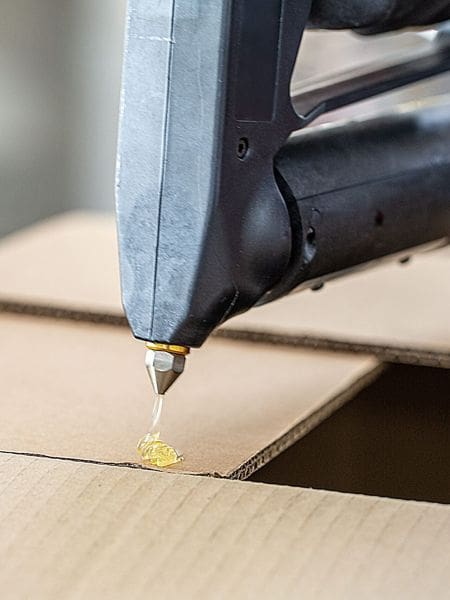 Handheld bulk hot melt sprayer applying hot melt adhesive to a cardboard carton